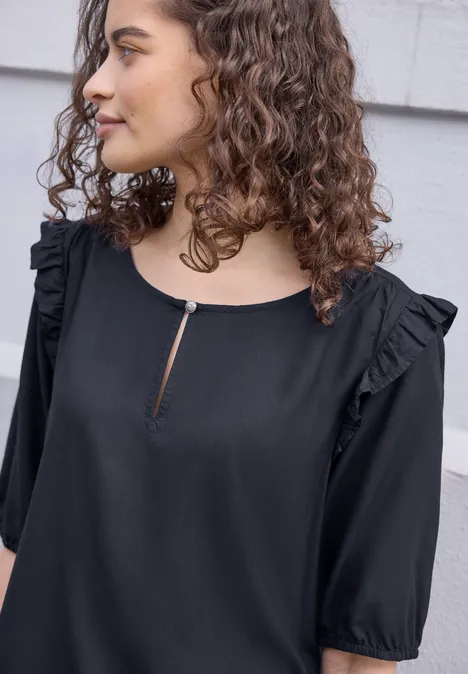 Kurzarm Bluse mit Volant-Details maritime blue Frau mit lockigem Haar trägt eine schwarze Bluse mit Rüschen an den Schultern und halblangen Ärmeln, Profilansicht.