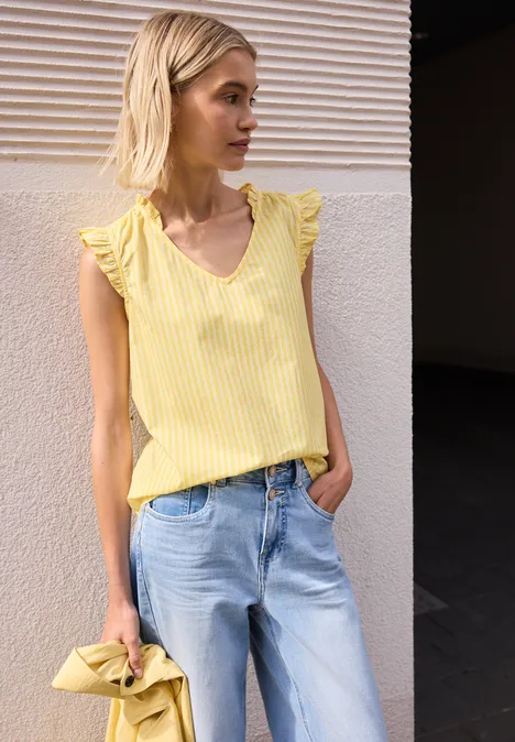 Frau lehnt an Wand und trägt gelbes Top mit Rüschenärmeln und helle Jeans.