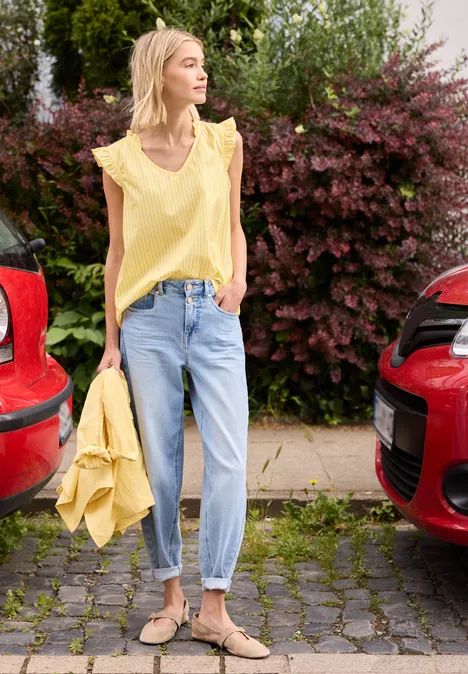Frau in schickem gelbem Top und Jeans steht zwischen zwei roten Autos, hält gelbe Jacke, sommerlich stylisch.