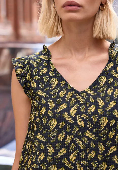Eine blonde Frau in schwarzem, gelb gemustertem Kleid mit V-Ausschnitt und Rüschenärmeln.