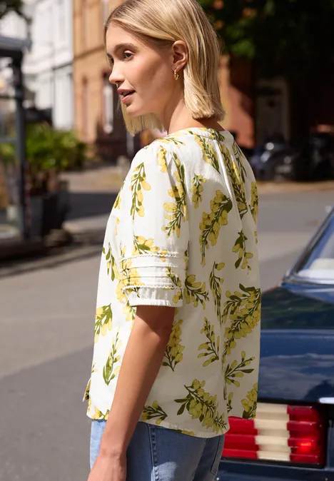 Zijaanzicht van persoon met wit bloesje met geel-groene bloemenprint naast auto op straat.