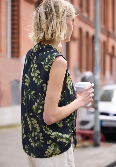 Glatte blonde Haare, lockeres schwarzes Top mit grünem Blattmuster, hält einen Coffee-to-go in der Stadt.