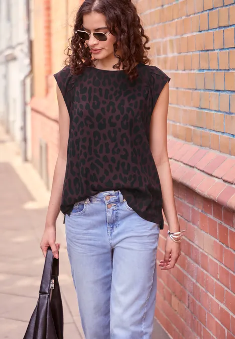 Eine Frau in Sonnenbrille, schwarzem Leo-Top und Jeans spaziert an einer Backsteinwand entlang.