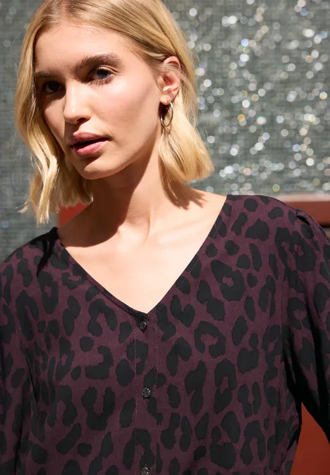 Frau mit kurzen blonden Haaren und Leopardenmuster-Kleid mit V-Ausschnitt vor glitzerndem Hintergrund.