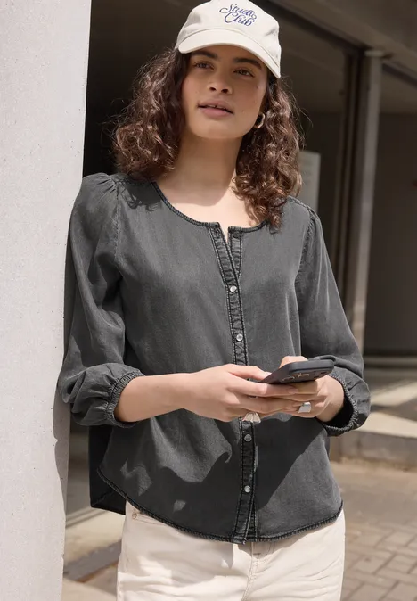 3/4-Arm Jeansbluse mit Rundhals tencel grey Frau in schwarzer Bluse und weißer Hose lehnt an einer Säule und hält ein Smartphone in der Hand.