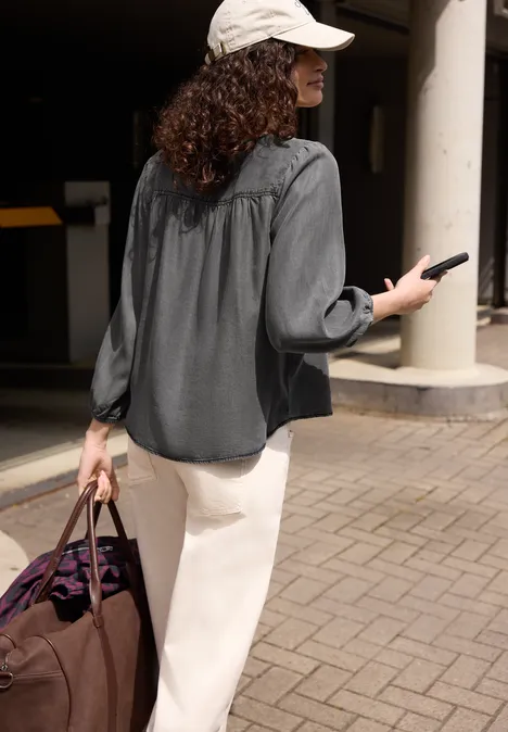 3/4-Arm Jeansbluse mit Rundhals tencel grey Frau mit weißem Hut, grauem Hemd und weißer Hose geht draußen mit Reisetasche und Smartphone.