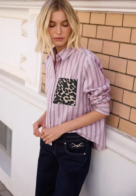 Femme en chemise rose à rayures avec poche léopard, pantalon foncé, devant un mur en briques.