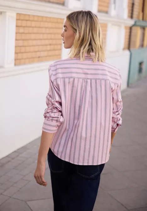 Femme de dos en chemise rayée rose et blanche, jean foncé, debout sur un trottoir devant un immeuble.