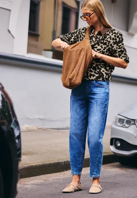 Frau in Jeans und Leopardenmuster-Bluse mit Tasche auf Gehweg zwischen parkenden Autos