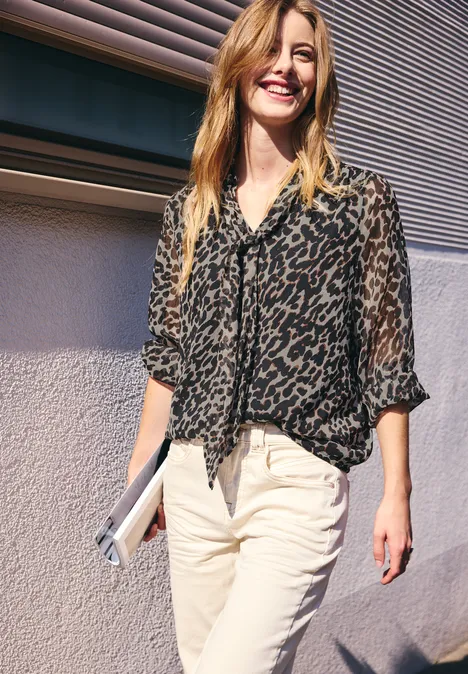 Femme souriante en chemisier léopard et pantalon clair, tient une pochette et marche près d'un mur.