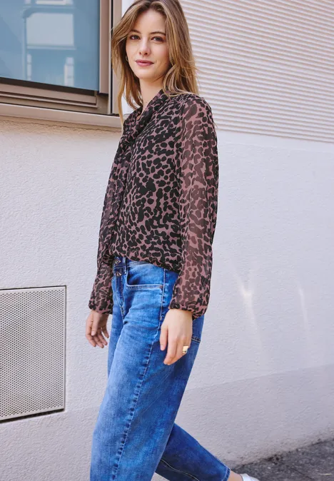 Jonge vrouw leunt nonchalant tegen een lichte muur, draagt een transparante luipaardblouse en blauwe jeans.