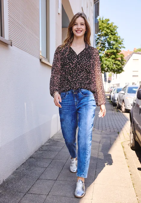 Een vrouw wandelt glimlachend over het trottoir, draagt een blouse, jeans en witte sneakers.