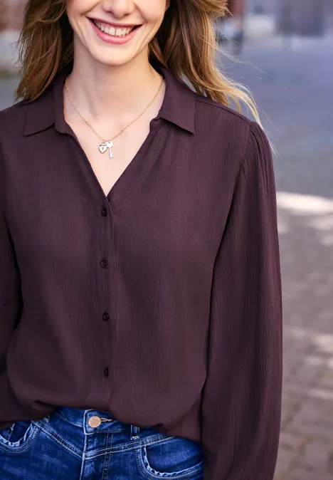 Vrouw in donkerpaarse blouse en spijkerbroek, glimlachend, met een fijne sleutelketting.