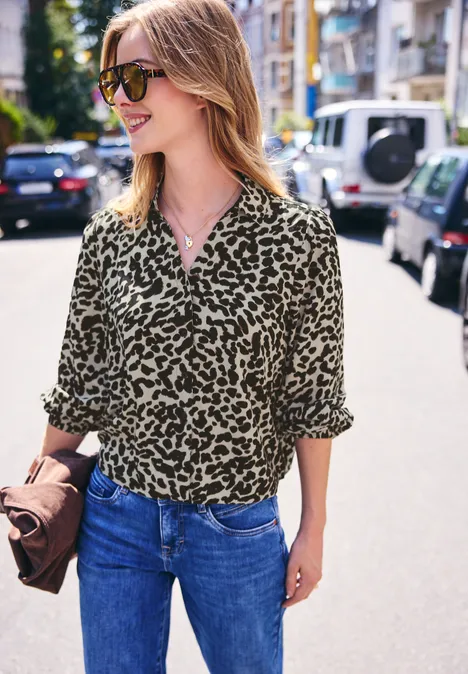 Vrolijke vrouw in luipaardblouse, spijkerbroek en zonnebril, clutch in de hand, wandelt in de zon.