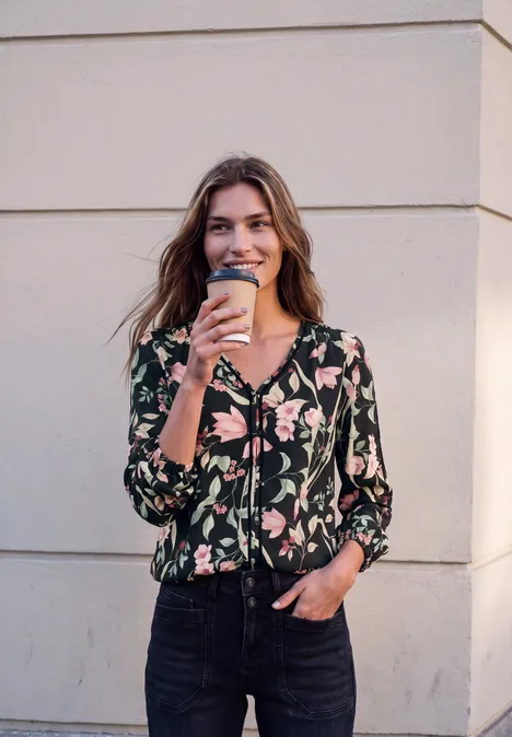 Eine lächelnde Frau mit floralem Oberteil und Jeans hält einen Kaffeebecher vor einer Wand.