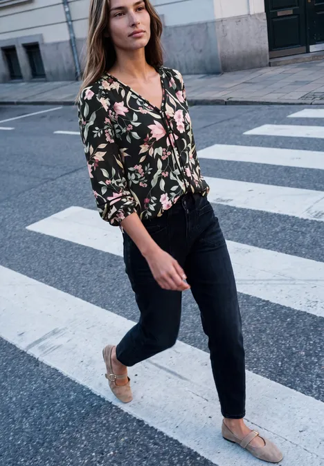 Eine junge Frau in einer geblümten Bluse und dunklen Jeans überquert einen Zebrastreifen.