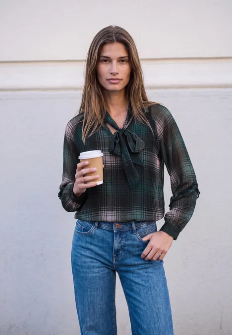 Eine Frau in kariertem Hemd und Jeans hält einen Kaffeebecher vor neutraler Wand.
