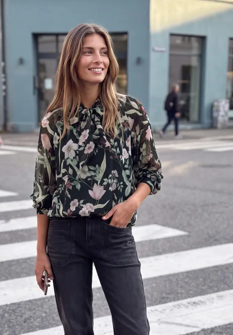 Frau mit floraler Bluse und Jeans steht lächelnd an einer Straßenkreuzung.