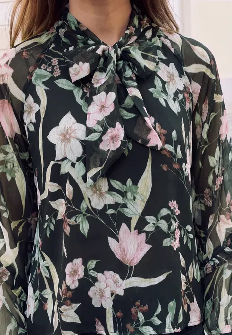 Schwarze Chiffonbluse mit rosafarbenen und weißen Blumen, grünen Blättern und Schleifenknoten am Kragen.