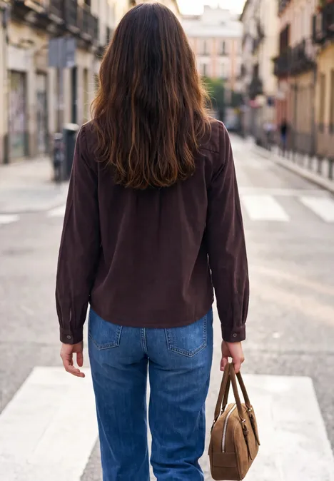 Eine Person geht von hinten gesehen über einen Zebrastreifen in einer Stadt, trägt Jeans, braune Jacke und Tasche.