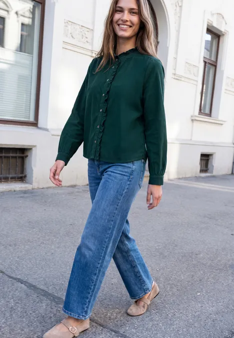 Eine junge Frau in grüner Bluse und blauen Jeans spaziert lächelnd eine ruhige Straße entlang.