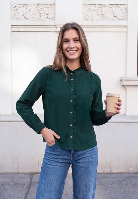 Junge Frau mit grünem Hemd und Jeans lächelt, hält einen Kaffeebecher vor weißer Hauswand.