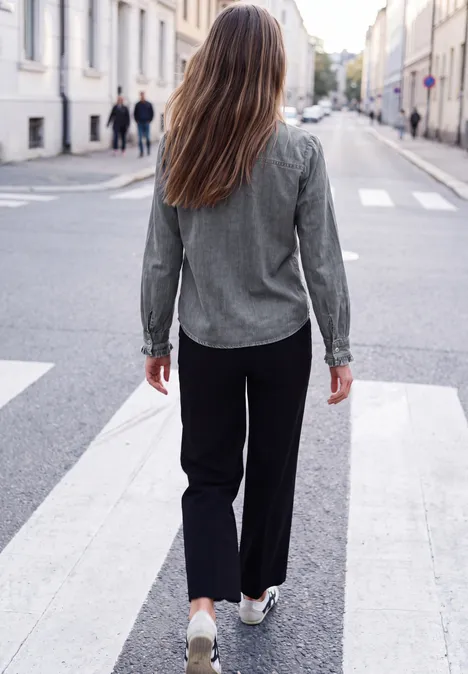 Langarm Denim Bluse mit Rüschen soft grey washed Eine Frau geht in legerer Kleidung über einen Zebrastreifen in einer ruhigen Stadtstraße.