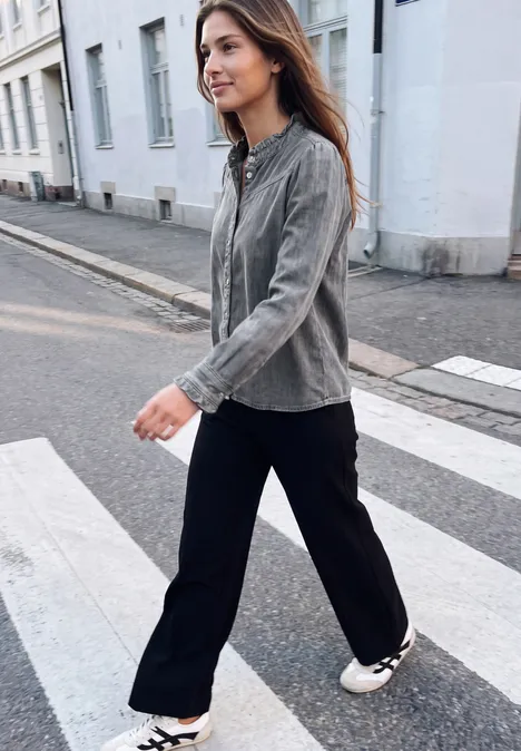 Langarm Denim Bluse mit Rüschen soft grey washed Eine junge Frau in grauer Jacke und schwarzen Hosen überquert einen Zebrastreifen in der Stadt.