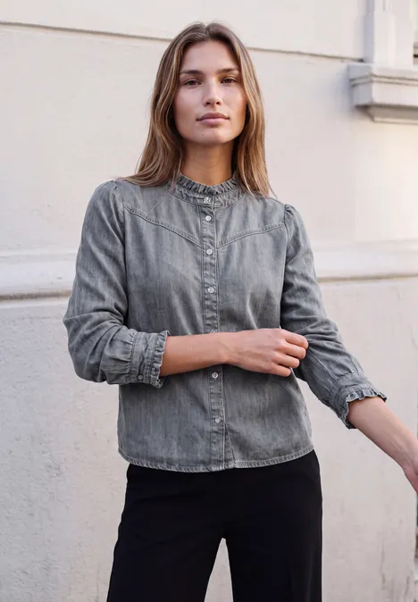 Langarm Denim Bluse mit Rüschen soft grey washed Eine Frau in grauem Jeanshemd und schwarzer Hose steht vor einer hellen Hauswand und schaut in die Kamera.