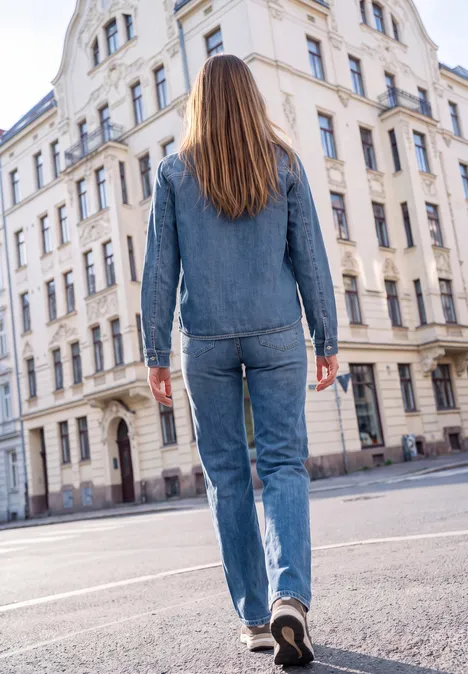 Eine Frau in Jeans-Outfit läuft auf einer Stadtstraße an einem alten Gebäude vorbei.