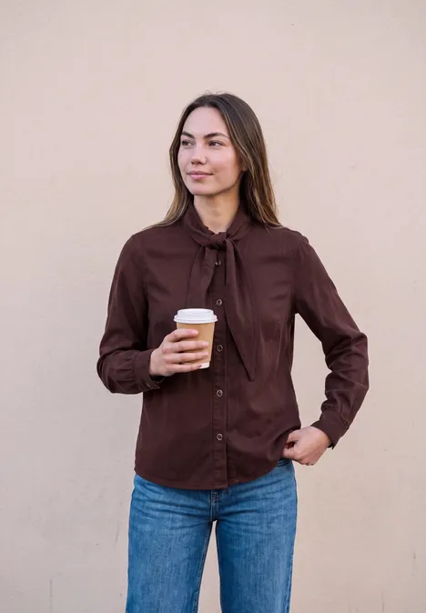 Eine Frau trägt braune Bluse und blaue Jeans, hält einen Kaffee und schaut zur Seite.