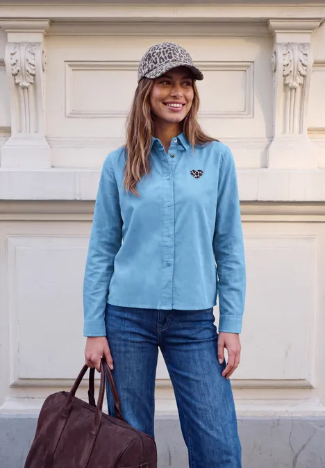 Chemisier en velours côtelé avec motif en forme de cœur loom blue Junge Frau vor cremefarbener Haustür, trägt hellblaues Hemd, Jeans, gemustertes Cap, hält braune Tasche, lächelt.
