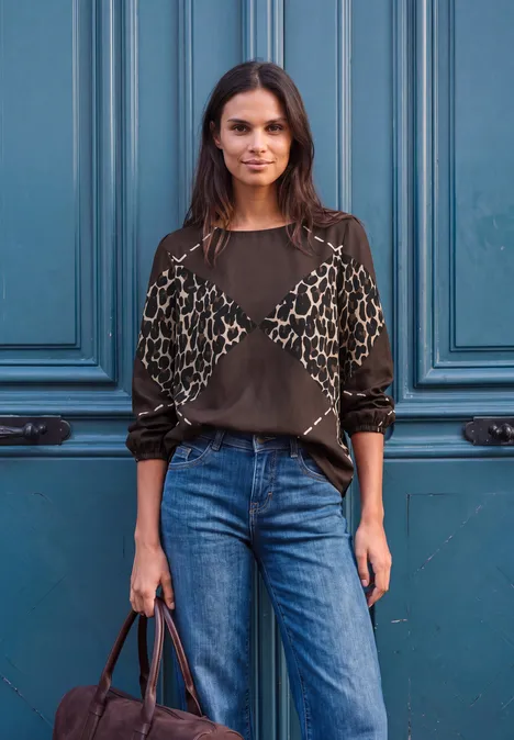 Eine Frau steht selbstbewusst vor einer blauen Tür, trägt Jeans und eine gemusterte Bluse.