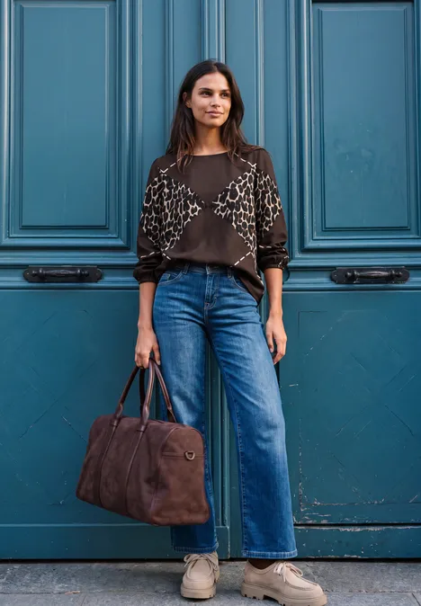 Eine Frau steht vor einer blauen Tür, trägt weite Jeans, gemusterten Pullover und hält eine braune Tasche.