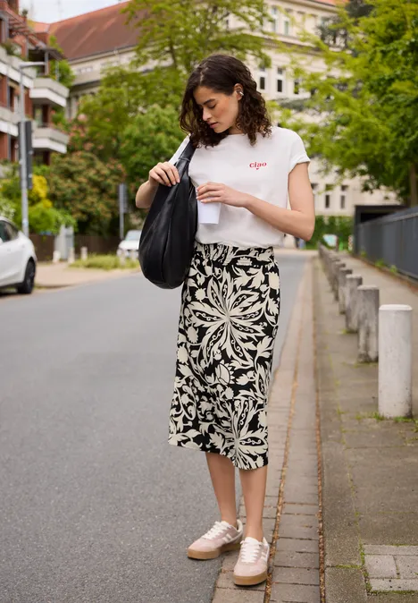 Midi-Rock mit Volants und Print Black Frau im weißen T-Shirt, schwarz-weißem Midirock und rosa Schuhen, mit schwarzer Tasche auf dem Gehweg einer Wohnstraße.