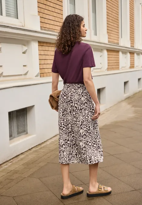 Midi-Rock mit Volants und Print very berry Frau von hinten in lila T-Shirt und gemustertem Midirock, mit Clutch und Sandalen auf Gehweg vor Haus.