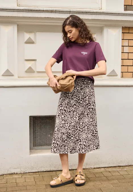 Midi-Rock mit Volants und Print very berry Frau in lila T-Shirt, gemustertem Midi-Rock und Sandalen steht mit Tasche vor Hausfassade.