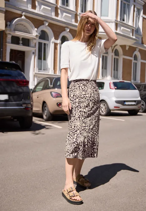 Frau in weißem T-Shirt und gemusterter Midirock mit Sandalen steht sonnig auf der Straße.