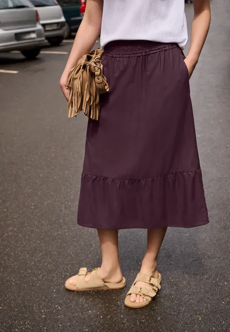 Midirock mit Volant und Elastikbund very berry Frau mit weißem Oberteil und pflaumenfarbenem Midirock, trägt Sandalen und braune Fransenhandtasche auf nasser Straße.