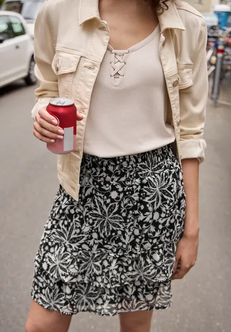 Knielange chiffonrok met volants Black Persoon in beige overhemd en gebloemde rok houdt buiten een koffie-to-go vast.