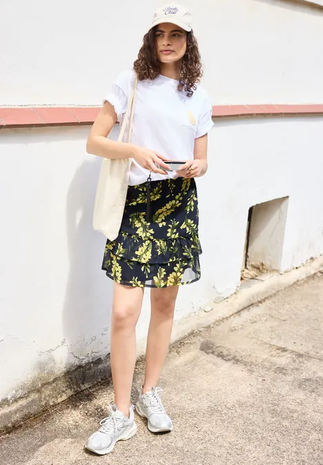 Knielanger Chiffon-Rock mit Print maritime blue Junge Frau in weißem T-Shirt, geblümtem Rock und Sneakers lehnt entspannt vor einer weißen Mauer.