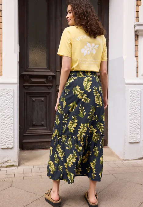 Eine Frau in gelbem T-Shirt und dunkelblauem Maxirock mit gelbem Blumenprint steht vor einem Hauseingang.