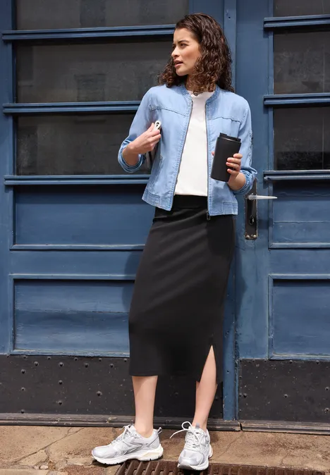 Midirock mit seitlichem Schlitz Black Frau steht vor blauem Tor, trägt Jeansjacke, weißes Shirt, schwarzen Midi-Rock, weiße Sneaker und hält Handy.