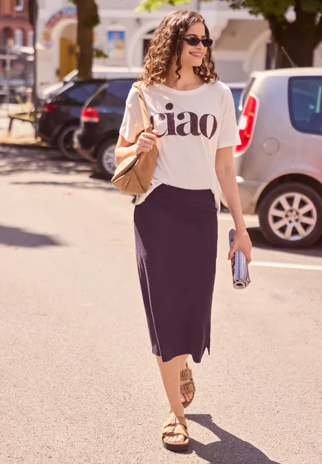 Vrouw in wit 'ciao'-shirt, zwarte splitrok, gouden sandalen en zonnebril wandelt in de felle zon op straat.