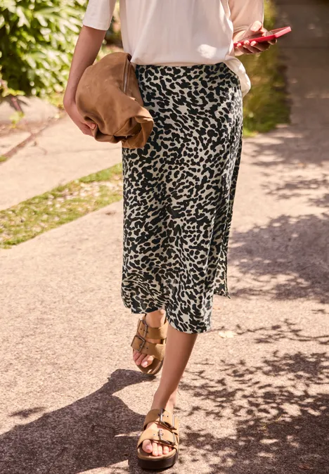 Frau in weißem Top und gemustertem Midirock, trägt goldene Sandalen und eine braune Tasche, geht auf sonnigem Gehweg.