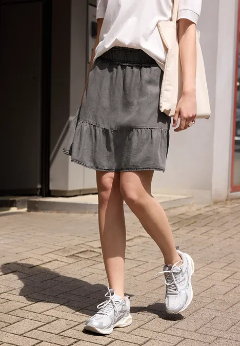 Vrouw in wit T-shirt, zwarte rok en witte sneakers met schoudertas loopt over een bestrate stoep.