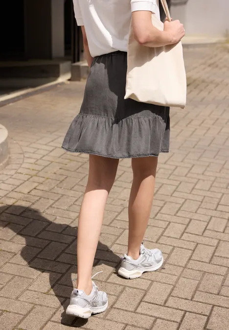 Vrouw van achteren in wit shirt, grijze rok en sneakers met een witte tas op een geplaveid pad.