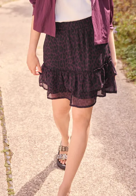 Frau in Leoparden-Rock und lila Jacke läuft auf gepflastertem Weg, trägt Sandalen mit Metallverschluss.