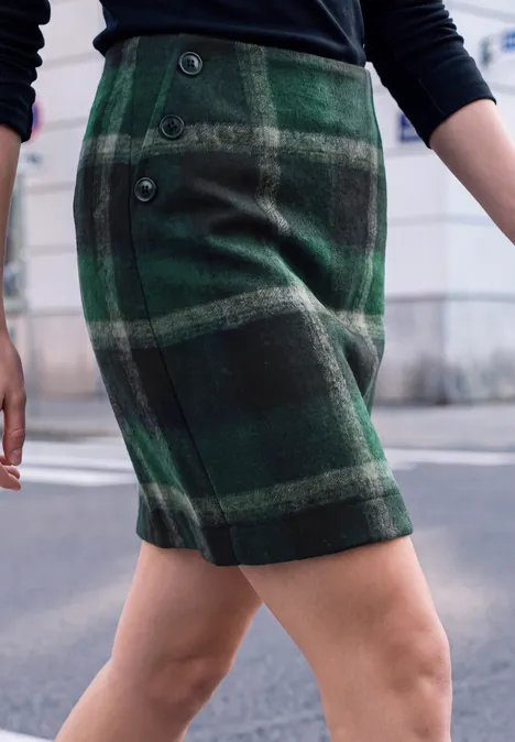 Iemand draagt een groene geruite minirok met zwart topje, alleen de benen en taille zijn zichtbaar.