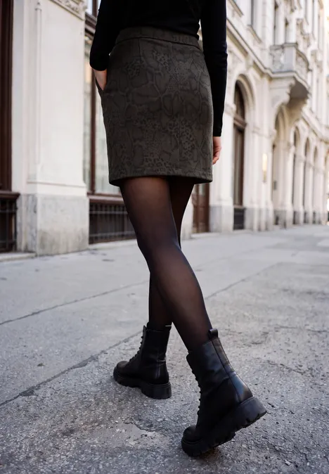 Minirock mit Snake-Print und Knopfdetails charred brown Eine Person läuft in kurzem Rock, schwarzer Strumpfhose und Boots eine Straße entlang, gesäumt von historischen Gebäuden.